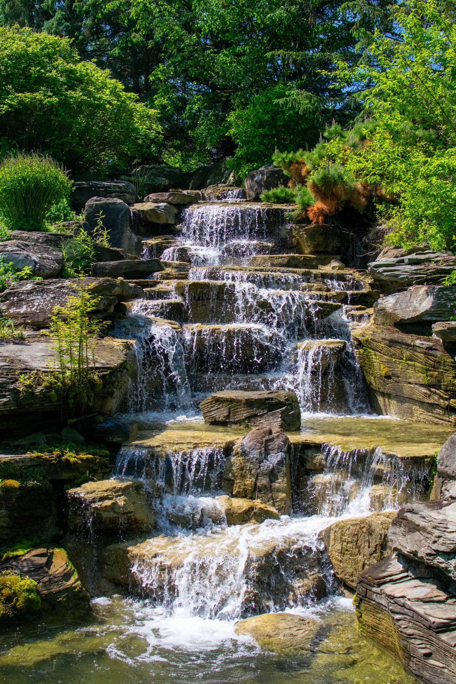 A tiered waterfall cascading over layered stone into a pool, surrounded by trees