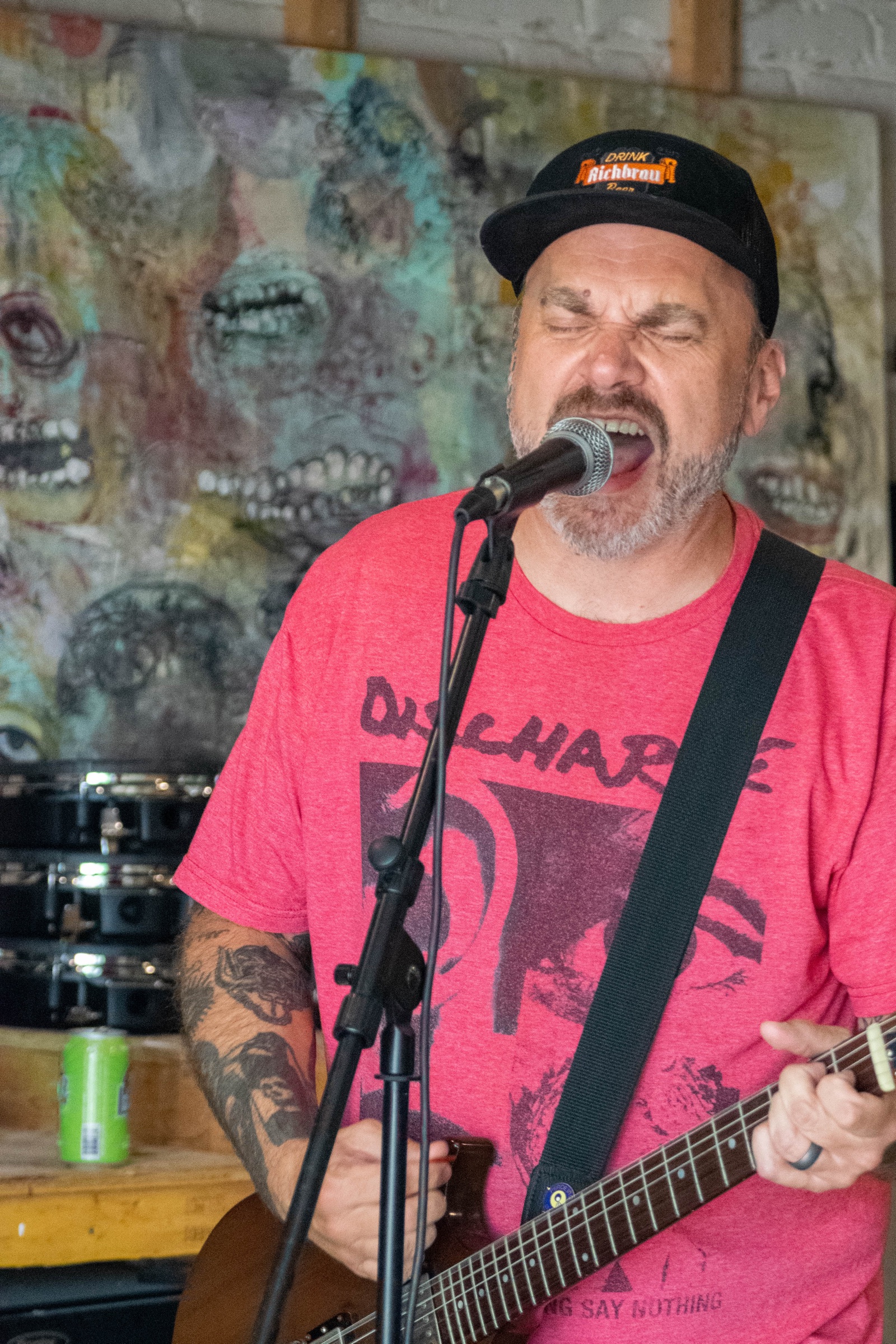 A guitarist in a red t-shirt and black cap singing into a microphone in front of a painted mural